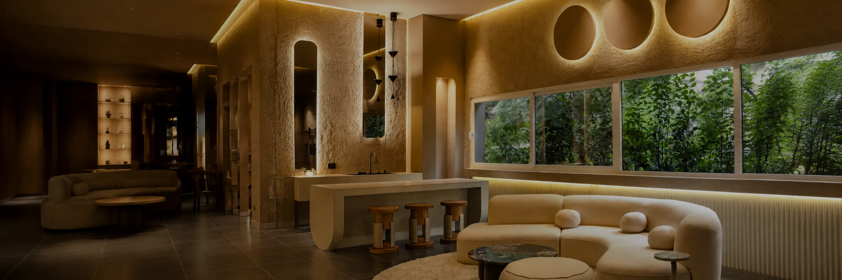 Sala de estar con iluminación LED arquitectónica: nichos circulares retroiluminados, cove lighting en cielorraso, sofá curvo y ventanal con vegetación